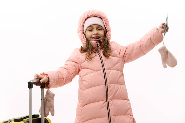 Radosna dziewczynka w ciepłych, puszystych ubraniach, jadąca na ferie zimowe, podnosząca ręce w podnieceniu, czekająca na nadchodzącą podróż lotniczą, pozująca z kartą pokładową i walizką na białym tle - Zdjęcie, obraz