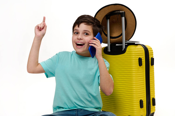 Teenage reiziger jongen in blauw turquoise t-shirt, met gele koffer, praten op mobiele telefoon, wijsvinger naar kopieerruimte op witte achtergrond, kijken naar camera, uiten van positieve emoties - Foto, afbeelding