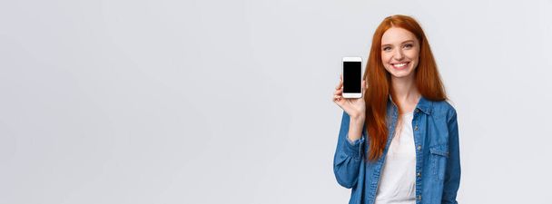 Wesoła, przyjazna rudowłosa kobieta sprawdza aplikację wodną, pozostaje nawodniona aplikacją na smartfona, pokazując ekran mobilny i uśmiechnięty zachwycony, stojący biały tło w ubraniu casual. - Zdjęcie, obraz