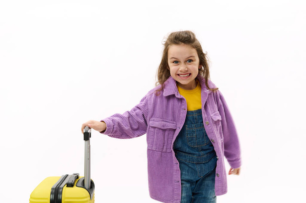 いたずら好きな旅行者の子供、紫のシャツとデニムの全体に身を包んだ少女は、休日のために行き、白い背景に黄色のスーツケースでポーズをとっています。海外旅行中の子供たち。旅行の旅 - 写真・画像