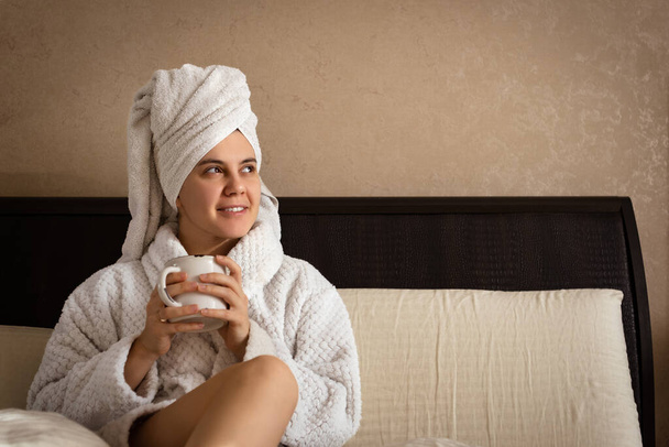 a woman sits on a bed wearing a white dressing gown, a towel covering her head, and a warm drink in her hands - Photo, Image
