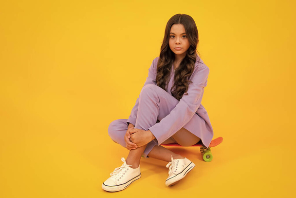 Teenager lifestyle. Beautiful and fashion teenager child girl in moder suit posing with a skateboard isolated on yellow background - Foto, immagini