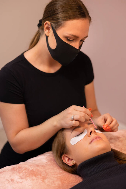 Cosmetologista feminina usando máscara protetora tratando jovem cliente para extensão de pestanas no spa de beleza durante a pandemia - Foto, Imagem