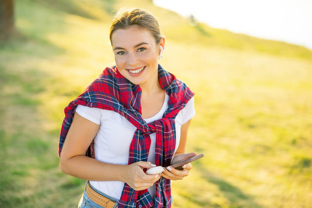 Uśmiechnięta dziewczyna słucha muzyki online za pomocą telefonu komórkowego i bezprzewodowych słuchawek w parku. Cyfrowy styl życia - Zdjęcie, obraz