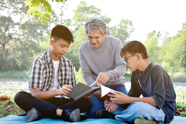 Asian elder and grandsons are spending time together in the park by teaching, reading and talking about topic in the book which they are interested in happily, soft  and selective focus on first boy. - Fotó, kép