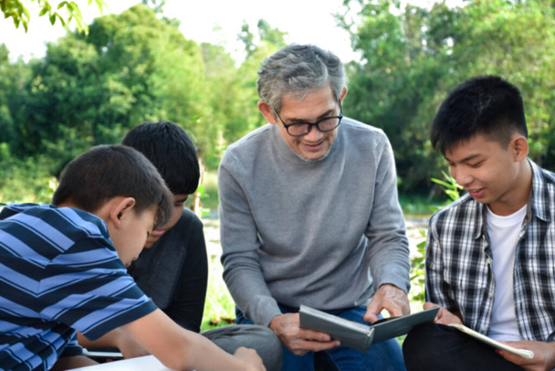 An elderly asian male is spending his spare time explaining the contents of the lesson and happily helping his two grandsons with their homework and school projects in his backyard, soft focus. - Foto, Imagem