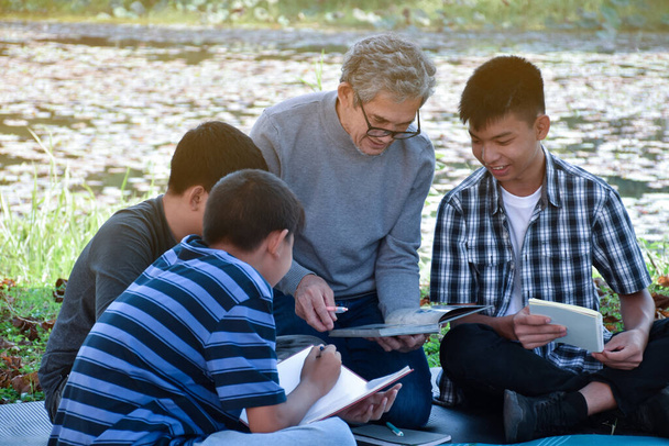 An elderly asian male is spending his spare time explaining the contents of the lesson and happily helping his two grandsons with their homework and school projects in his backyard, soft focus. - Foto, Bild