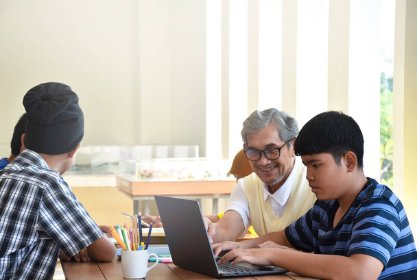 Asian boys are sitting infront of asian elderly teacher inside the room to ask and to do the school project work and listening to the elderly teacher about their project work, soft and selective focus - Foto, imagen