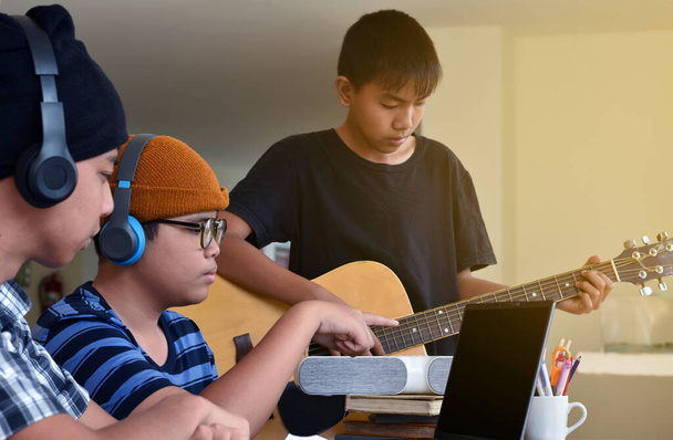 Group of young asian teenagers sitting together inside the room working and practising school project about the music subject through laptop and playing quitar, too, soft and selective focus. - Fotografie, Obrázek