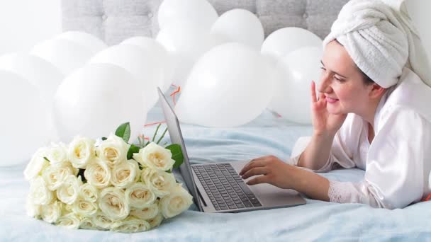 Woman in white clothes and towel on her head lying on a bed with white color balloons and bouquet of roses. Woman works at home, using her laptop, computer. Birthday, wedding. Copy space. Preparation. - Felvétel, videó