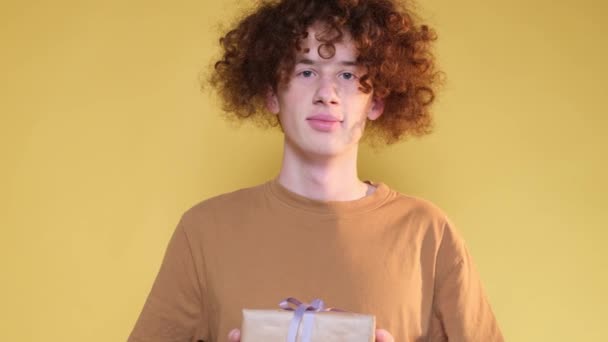 赤い髪のハンサムな男が贈り物を持っている黄色の紙に包まれ、青いリボンで結ばれています。若い男が贈り物を示す - 映像、動画