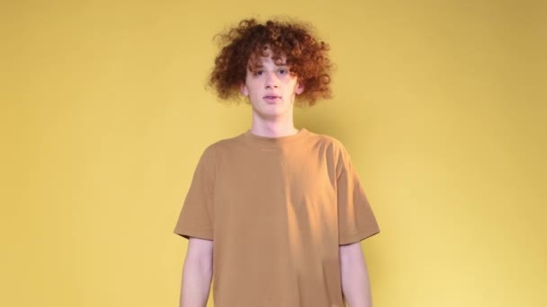 Surprised curly-haired guy spreads his hands and looks at the camera on a yellow background in the studio. Red-haired teenager. - Metraje, vídeo