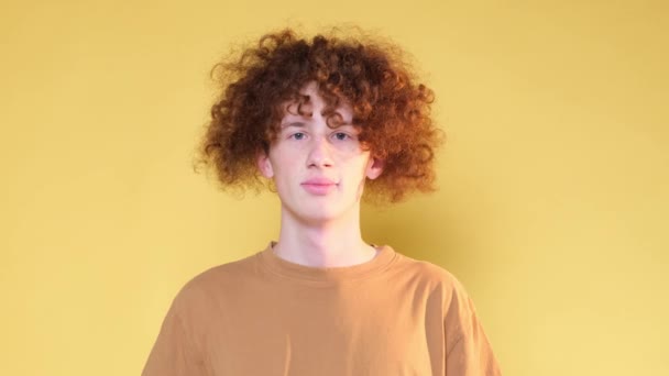 Πορτρέτο του κοκκινομάλλη σγουρομάλλη έφηβο δείχνει αντίχειρες επάνω. Ένας γελαστός έφηβος με σιδεράκια στα δόντια, απομονωμένος σε κίτρινο φόντο στο στούντιο. Βίντεο 4k - Πλάνα, βίντεο