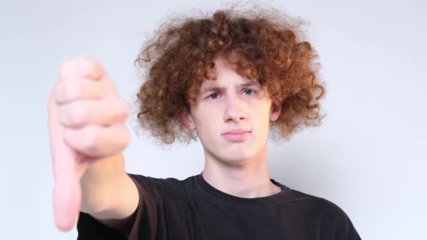 Portrait of a curly man with a sign of dislike. Young guy expressing displeasure by showing thumb down, on gray background in studio. Video 4k - Filmagem, Vídeo