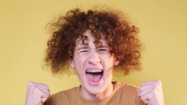 Curly man shows crazy joy, isolated on yellow background in studio. Joyful emotions of a young guy. The concept of happiness and victory - Metraje, vídeo