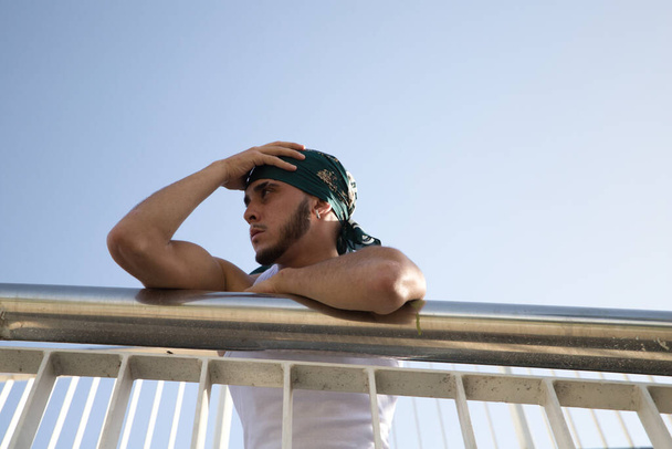 Portrait of a young handsome man dressed in jeans and white t-shirt and green headscarf. The young man is leaning on railing and makes different expressions. Street fashion and modern youth culture. - Foto, Imagen