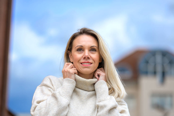 Mulher loira de meia idade atraente segurando sua gola alta com as mãos em uma cidade contra um belo céu azul em visão de baixo ângulo - Foto, Imagem