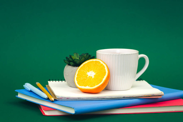 Notebooks, a cup of tea and an orange on a green background, the concept of a school snack. - Photo, Image