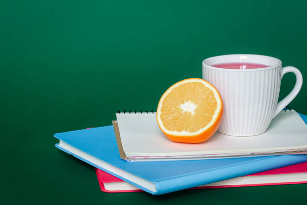 Notebooks, a cup of tea and an orange on a green background, the concept of a school snack, copy space. - Photo, Image