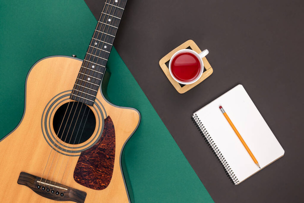 Επίπεδη lay, ακουστική κιθάρα, φλιτζάνι τσάι, και σημειωματάριο στο φόντο χρώμα. - Φωτογραφία, εικόνα