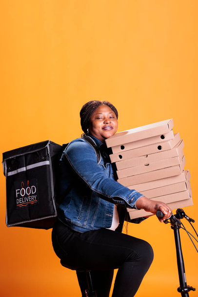 Glimlachende afhaalmaaltijd vrouw met kartonnen doos stapel vol met pizza leveren afhaalmaaltijden bestelling aan de klant tijdens het rijden fiets. Persoon die in studio over gele achtergrond staat - Foto, afbeelding