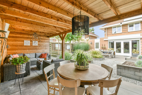 un patio con sillas, mesas y plantas en la mesa frente a una sala de estar al aire libre que tiene paredes con paneles de madera - Foto, Imagen