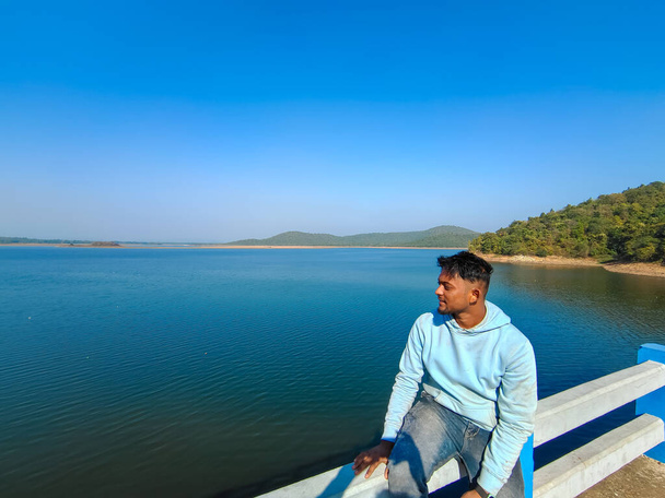 A young boy sitting on the bridge and looking at blue river, lake with blue sky scenery. Holiday concept  - Photo, image
