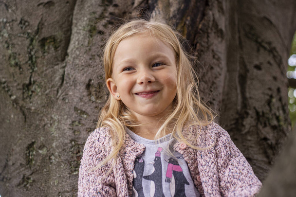 Smiling child girl is playing sitting high on a tree in the park. - Photo, Image