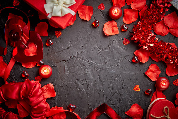 Понятие Дня влюбленных. Красные лепестки роз, свечи, аксессуары для свиданий, коробочные подарки, сердечки, блёстки на чёрном фоне, рамка композиции, вид сверху. Макет для поздравительных открыток
 - Фото, изображение