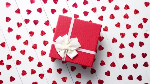 Composition décoration Saint-Valentin. Cadeau en boîte placé sur des paillettes rouges en forme de coeur sur une table en bois blanc. Fond romantique. Couché plat, vue du dessus
. - Séquence, vidéo