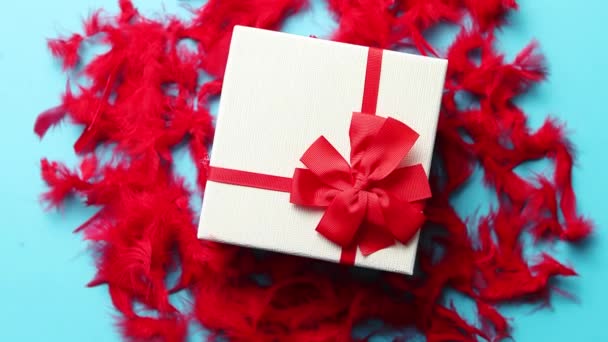 Caja con un regalo, atada con una cinta colocada sobre plumas rojas. Vista superior, primer plano, plano, fondo azul aislado. Concepto para San Valentín
 - Imágenes, Vídeo