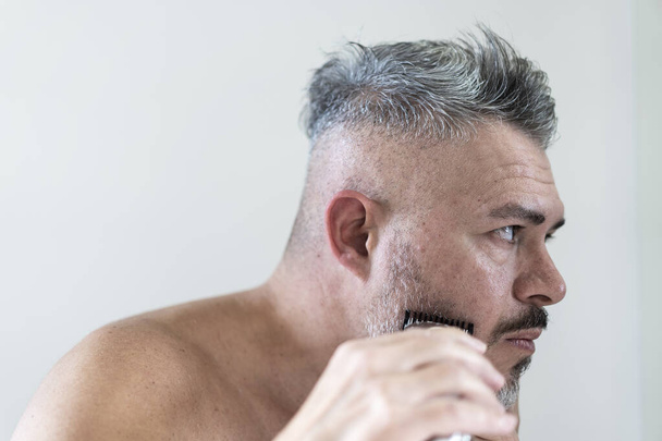 Jóképű ősz szakállas férfi vágja a szakállát egy trimmer - középkorú férfi nézi magát a tükör és vágás a szakáll a fürdőszobában - Szépség, higiénia, borotválkozás, ápolás koncepció - Fotó, kép
