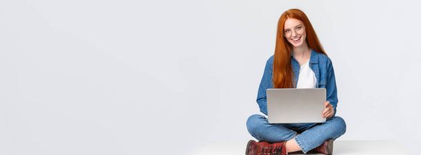 Neşeli, küstah, kızıl saçlı kız öğrenci, gözlüklü üniversiteli kız dizüstü bilgisayarla bacak bacak üstüne atmış, serbest çalışıyor, uzaktan kumandalı beyaz arka plan üzerinde projeyi hazırlıyor.. - Fotoğraf, Görsel