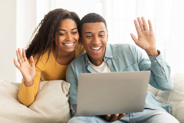 技術は、リモートで会う。100万人のアフリカ系アメリカ人の夫婦がノートパソコンを見てビデオ通話をし、光のリビングルームのインテリアに手を振ってください。挨拶や挨拶や家でのコミュニケーション - 写真・画像