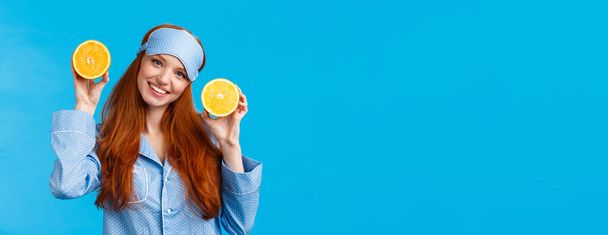 Girl suggesting fruit for breakfast. Cute and tender redhead woman in sleep mask, pyjama, tilt head and holding two slice orange, smiling, prepare juice, eating healthy morning, blue background. - Photo, Image