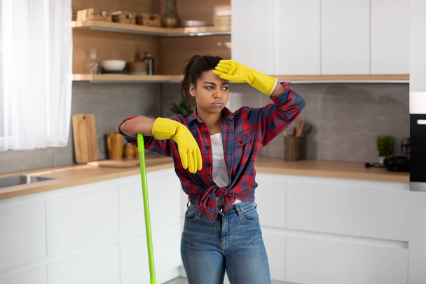 Unhappy millennial african american housewife in rubber gloves wipes her forehead and mop floor in minimalist kitchen interior. Tired of cleaning house alone, a lot of work on weekends and service - Photo, image