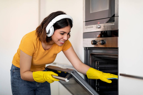 Mutfakta temizlik yaparken fırın temizleyen mutlu genç Arap kadın, neşeli Orta Doğulu ev hanımı lastik eldivenler giyip evi topluyor ve favori müziğini dinliyor, yakın çekim - Fotoğraf, Görsel