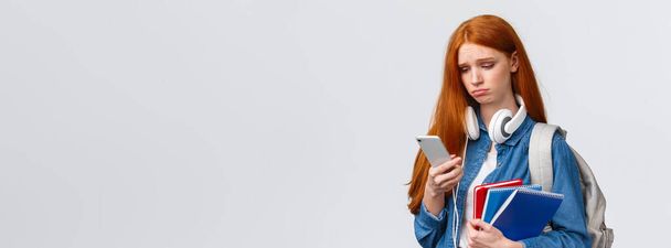 Silly sad and gloomy pouting girl with red foxy hair, frowning sighing and looking with regret smartphone screen, reading troublesome message, feeling bad, stand with backpack and notebooks. - Photo, Image
