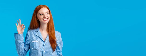 Dreamy and creative cute redhead woman feeling happy and relieved, wearing nightwear, showing recommend, okay gesture, approving new hygiene product, standing blue background. - Photo, Image
