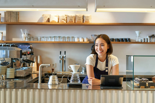 Sonriendo amigable chica asiática barista, usando delantal, trabajando en el mostrador, la elaboración de café filtro, la gestión de la tienda de café. - Foto, Imagen