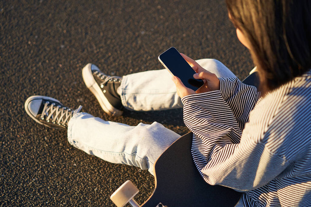 Κοντινό πλάνο του skater έφηβος κορίτσι κάθεται στο skateboard, πληκτρολογεί μήνυμα στο smartphone, κοιτάζει οθόνη του κινητού τηλεφώνου. - Φωτογραφία, εικόνα