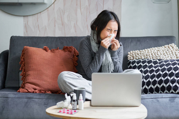 家にいる若いアジアの女性は、体調が悪く、風邪をひいたまま、ラップトップに座って、コンピュータで映画を見て、くしゃみをする. - 写真・画像