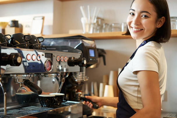 Χαμογελαστή Ασιάτισσα που δουλεύει στο καφέ, χρησιμοποιεί καφετιέρα για εσπρέσο, παραγγελίες καπουτσίνο, στέκεται πίσω από πάγκο με μπλε ποδιά, κοιτάζοντας την κάμερα. - Φωτογραφία, εικόνα