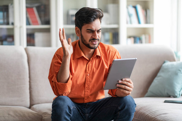 Happy cheery smiling handsome middle eastern millennial man in casual outfit sitting on couch, waving at digital pad screen, guy having online party with friends, home interior, copy space - Foto, Imagen