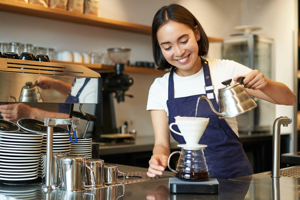 Χαριτωμένο μελαχρινή κοπέλα barista, καφέ προσωπικό ρίχνει νερό από το βραστήρα και ζυθοποιίας φίλτρο καφέ πίσω από τον πάγκο, την προετοιμασία της παραγγελίας. - Φωτογραφία, εικόνα