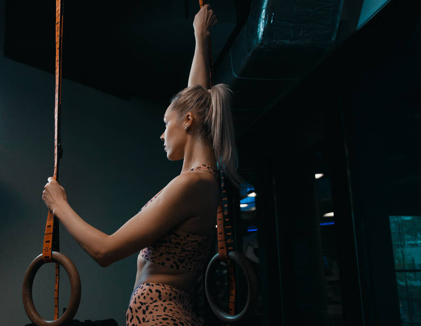 Young fit girl pulling up on a gimnastic rings. Muscular back of a girl Crossfit athlete in black sportswear is hanging on gymnastic rings. Caucasian woman with long blond hair training olympic ring. - Foto, imagen