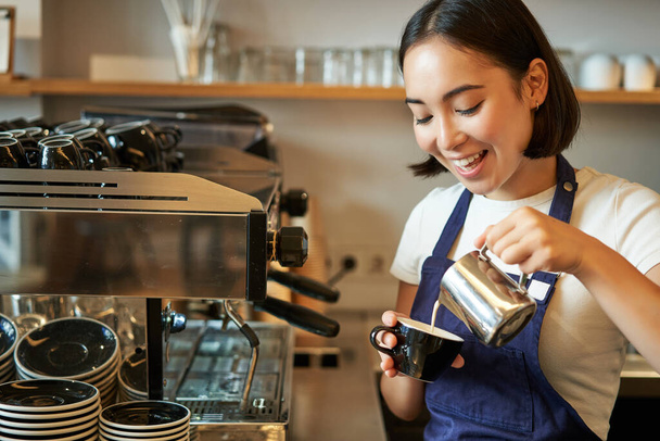 Close up di carino asiatico barista ragazza making cappuccino, doing latte art in cup with steamed milk, standing in coffee shop behind counter. - Foto, immagini