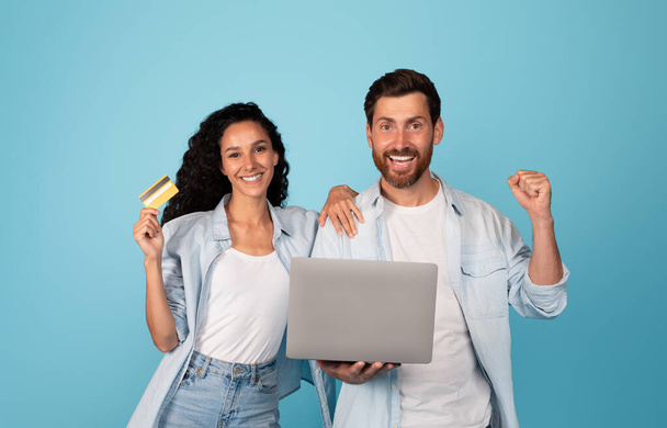 Smiling young european couple with credit card and laptop make gesture of victory and success, great purchase, sale, isolated on blue background, studio. Online shopping for shopaholics and cashback - Photo, image