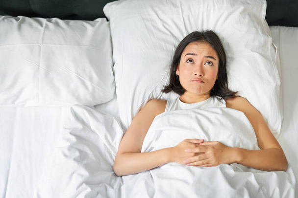 Yatağında yatan ve omuz silken Asyalı bir kızın portresi. Şaşırmış görünüyor. Uyumadan önce çok düşünüyor.. - Fotoğraf, Görsel