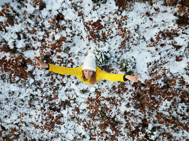 彼女の腕で雪の性質に立っている鮮やかな黄色のセーターで若い女性のオーバーヘッドビューは高いと彼女の顔に笑顔を上げ. - 写真・画像